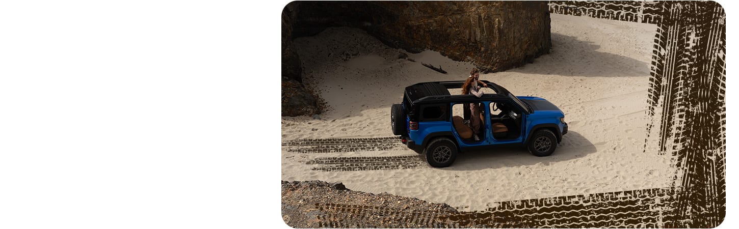 Un Jeep Recon Moab 2026 azul sin las puertas, estacionado en la playa con una pareja de pie en la fila de atrás mirando a través del techo corredizo.