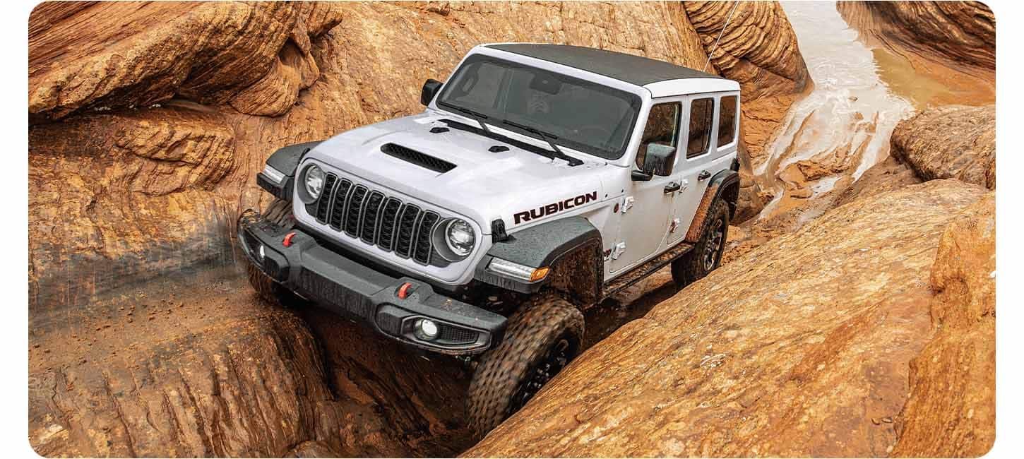 Un Jeep Wrangler Rubicon 2026 blanco que avanza lentamente por una ruta todoterreno extrema, con las ruedas delanteras y traseras del lado del conductor encajadas en un surco y las del lado del copiloto sobre un saliente de piedra elevado.