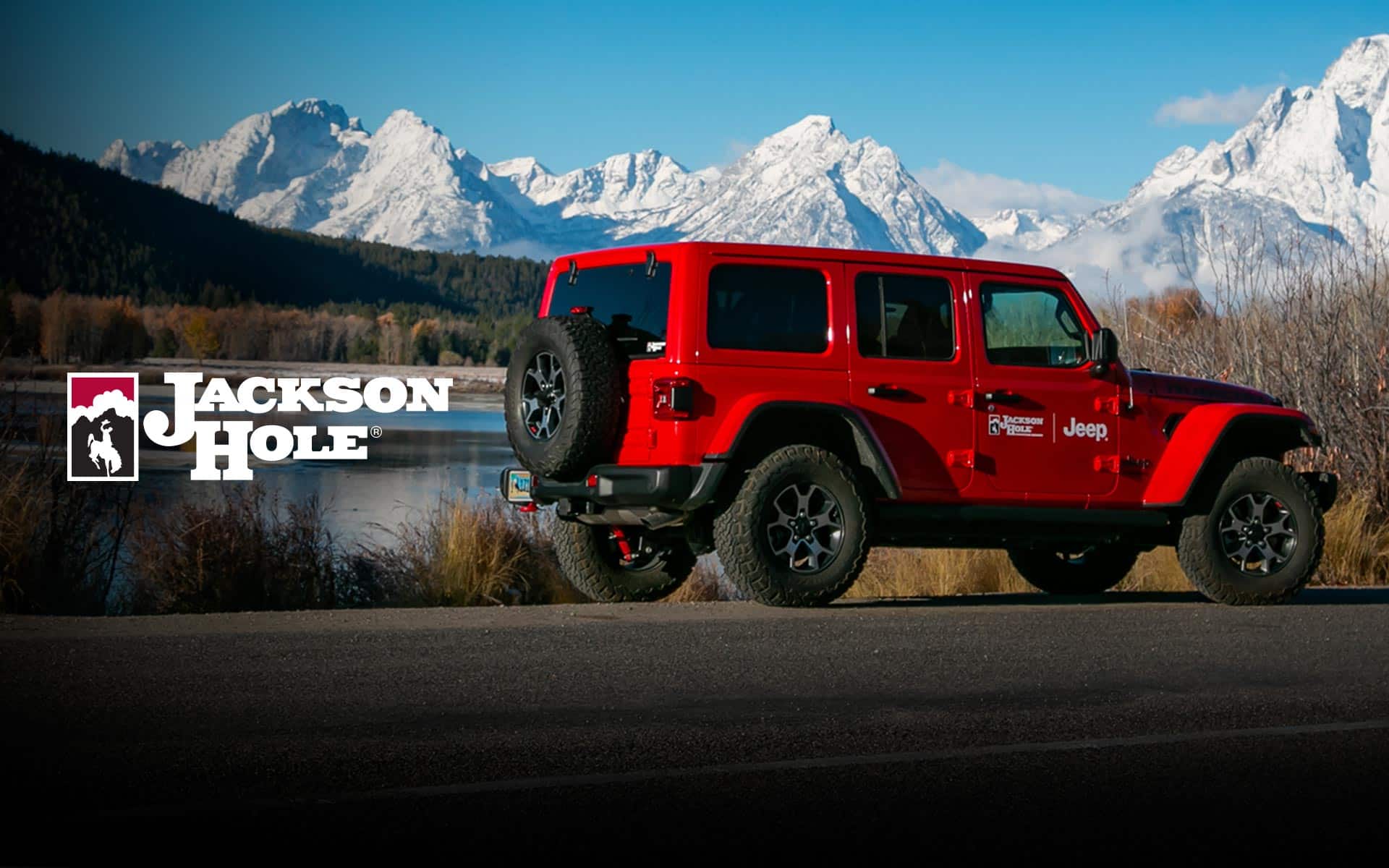 A profile of a red Jeep Wrangler Rubicon with Jackson Hole logo on the passenger-side door, parked beside a lake, with mountains in the background. Jackson Hole.
