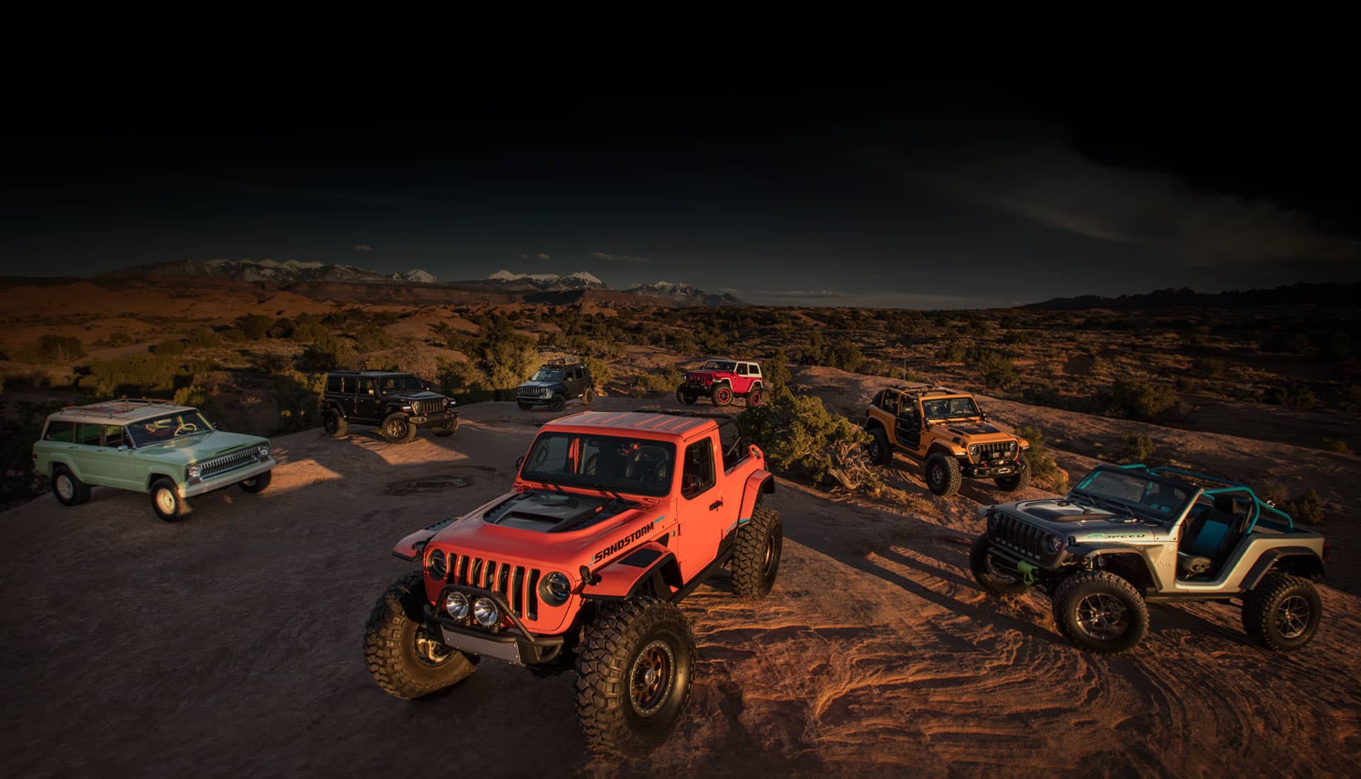 Una combinación de siete vehículos conceptuales de la marca Jeep estacionados en una meseta rocosa en las montañas, por la noche, iluminados con luz artificial.