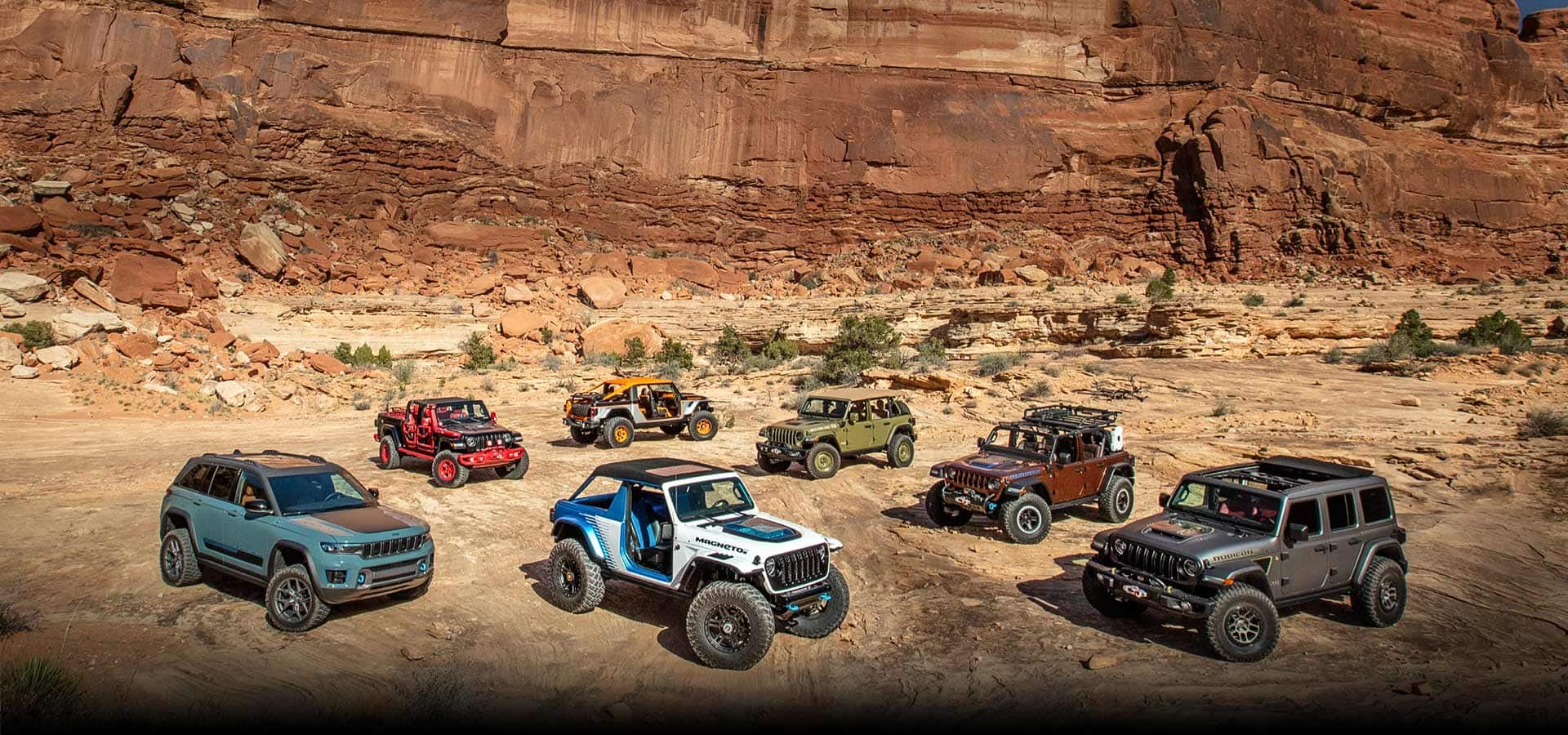Seven Jeep Brand concept vehicles parked on a sandy plain with a red rock cliff rising behind them.