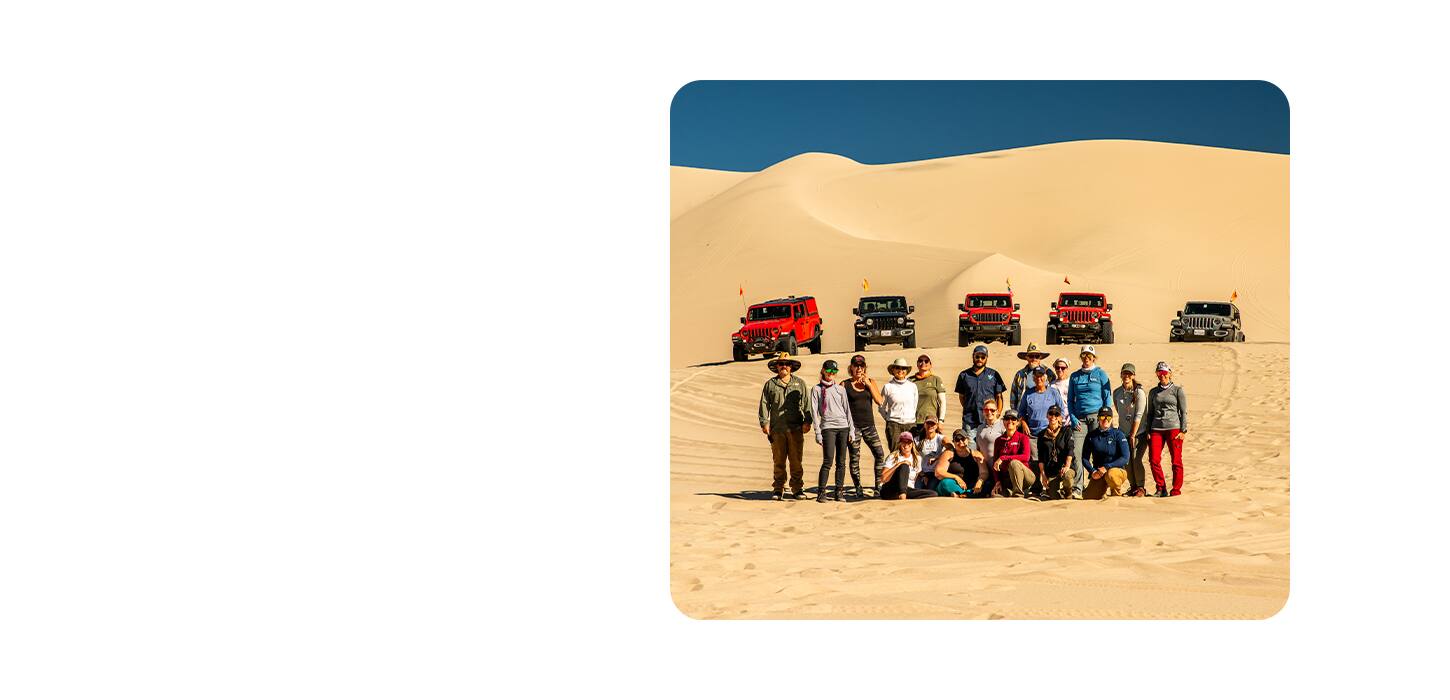 Un evento de la marca Jeep en el desierto, con un grupo de propietarios de vehículos de la marca Jeep en primer plano y cinco vehículos de la marca Jeep de fondo.