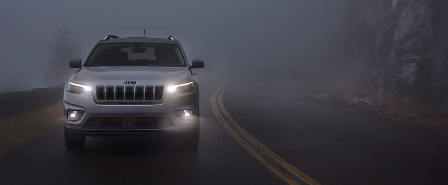 The 2021 Jeep Cherokee Altitude being driven on a winding, foggy road at night with headlamps and fog lamps lit.