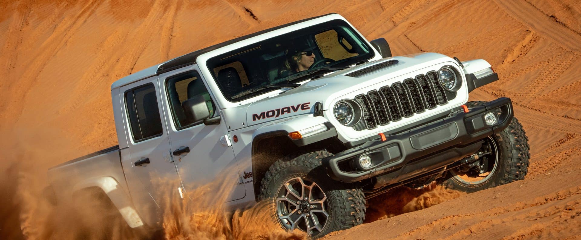 Un Jeep Gladiator Mojave blanco levantando arena mientras sube por una duna en el desierto.