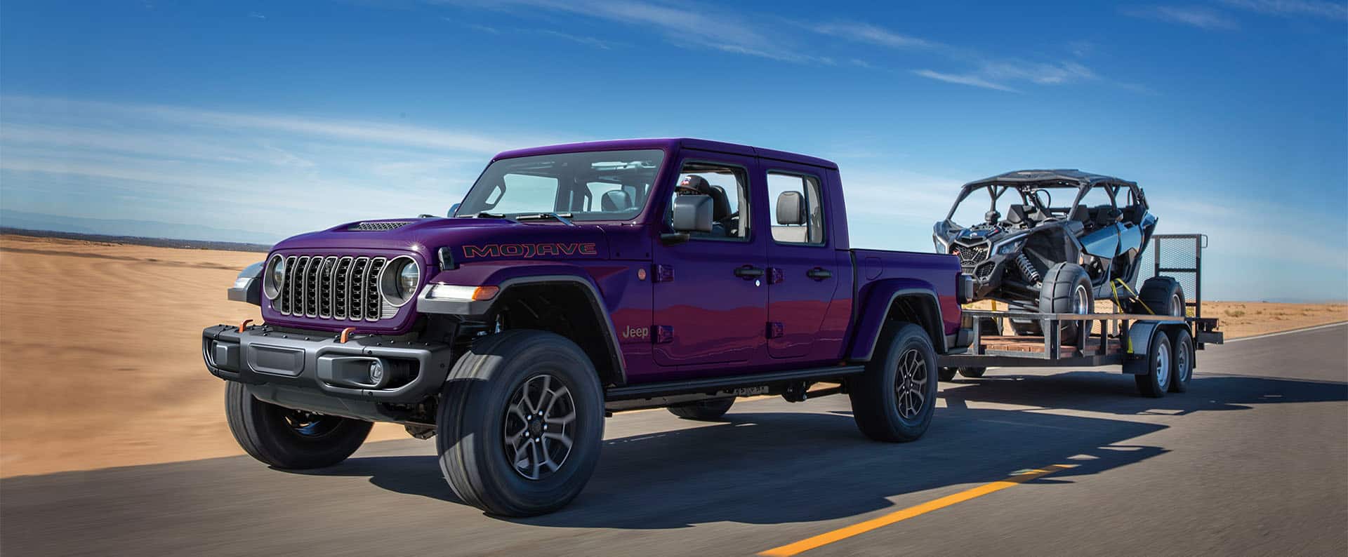 A dark purple 2026 Jeep Gladiator Mojave X traveling on the highway, towing an ATV.