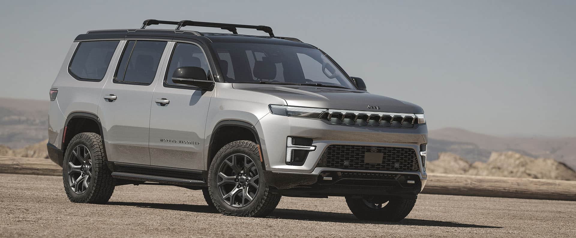 A passenger-side front angle of a silver 2026 Grand Wagoneer Upland, parked on a clearing, with mountains in the background.