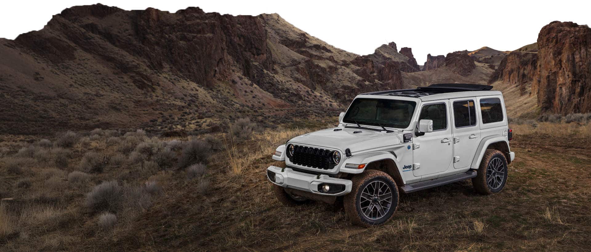 Un Jeep Wrangler Sahara 4xe 2025 blanco estacionado en un claro, fuera de la carretera, entre colinas y afloramientos.