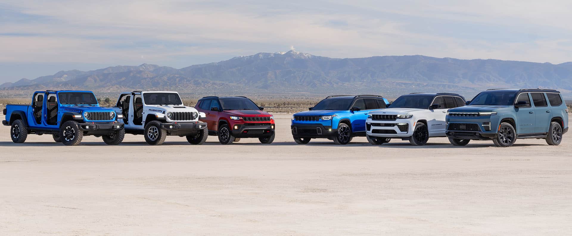 A lineup of six Jeep Brand vehicles parked on a grassy clearing with mountains in the background. From left to right: a gray 2026 Gladiator Mojave X, a blue 2026 Wrangler Rubicon X, an orange 2026 Compass Limited, a blue 2026 Cherokee Overland, a red 2026 Grand Cherokee Summit and a silver 2026 Grand Wagoneer Summit Reserve.