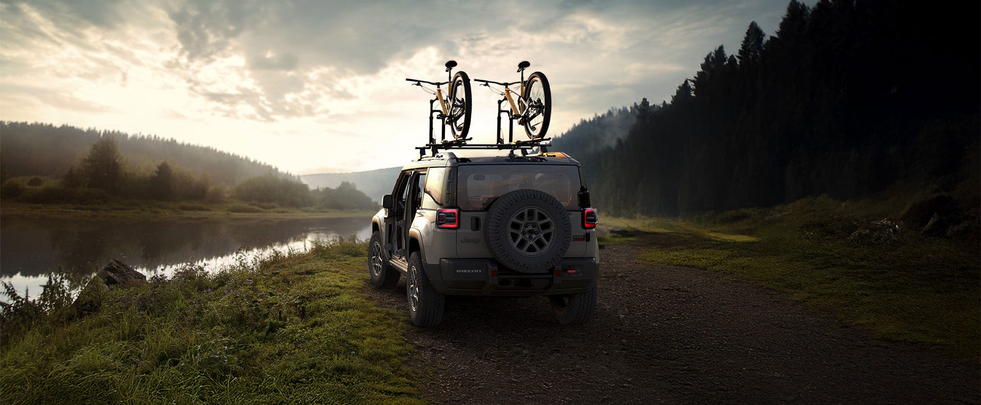 La parte trasera de un Jeep Recon 2026 verde oliva con las puertas desmontadas y dos bicicletas amarradas a la parrilla portaequipaje del techo, estacionado en un sendero cubierto de césped, con bosques de montaña de fondo.