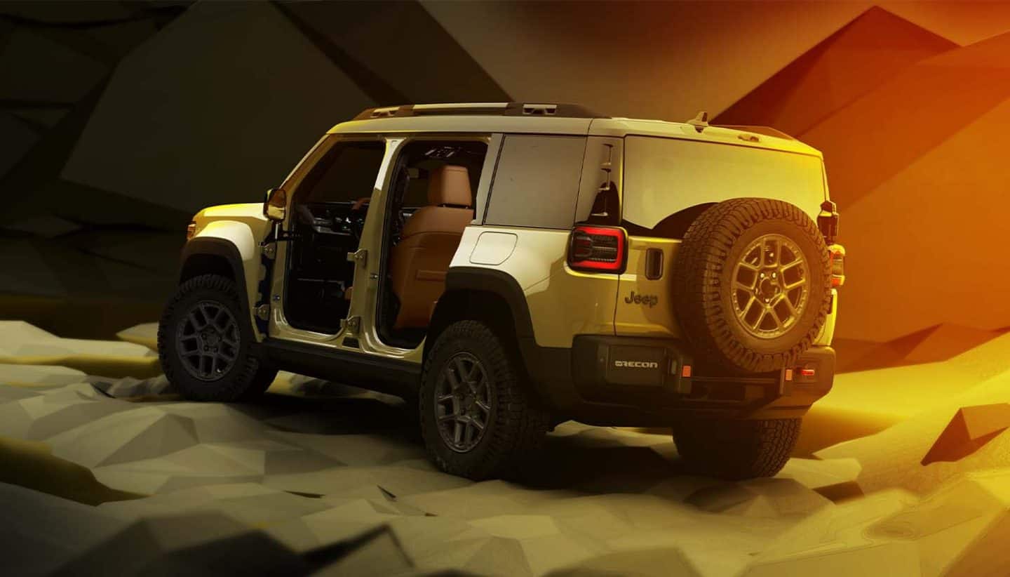 Display A driver-side rear angle of a 2026 Jeep Recon with its doors removed, parked in a studio with an abstract background and shaded lighting that goes from olive green to golden yellow and orange.