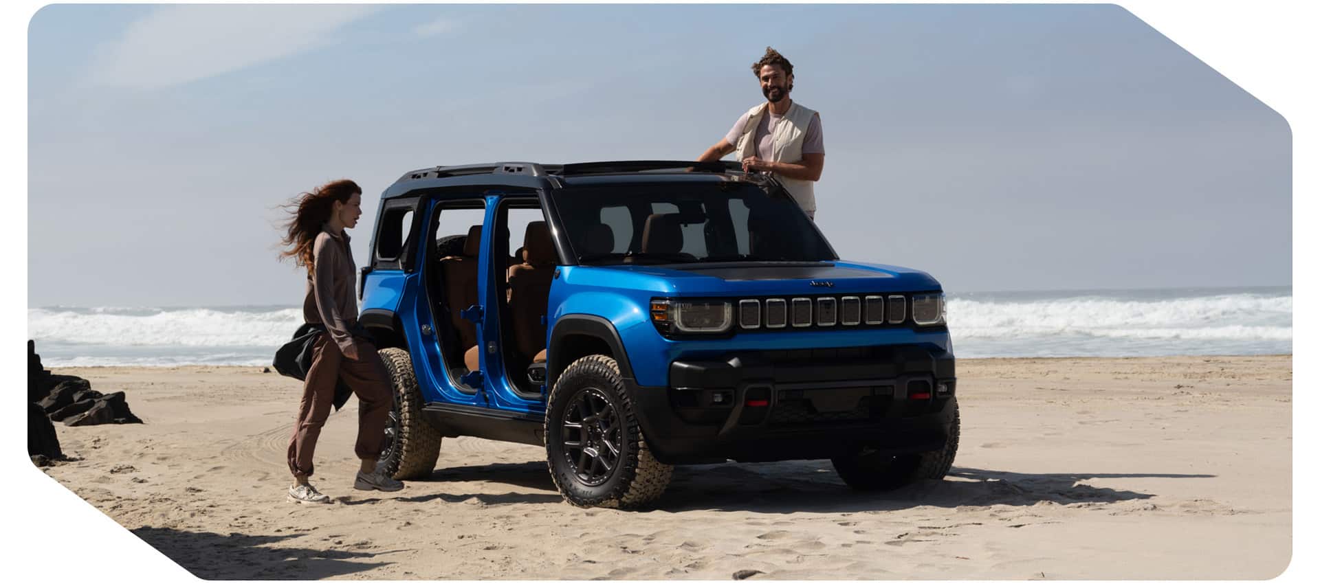 Una toma frontal del lado del pasajero de un Jeep Recon 2026 azul con las puertas removidas, estacionado en una playa de arena con una mujer y un hombre a cada lado del SUV.