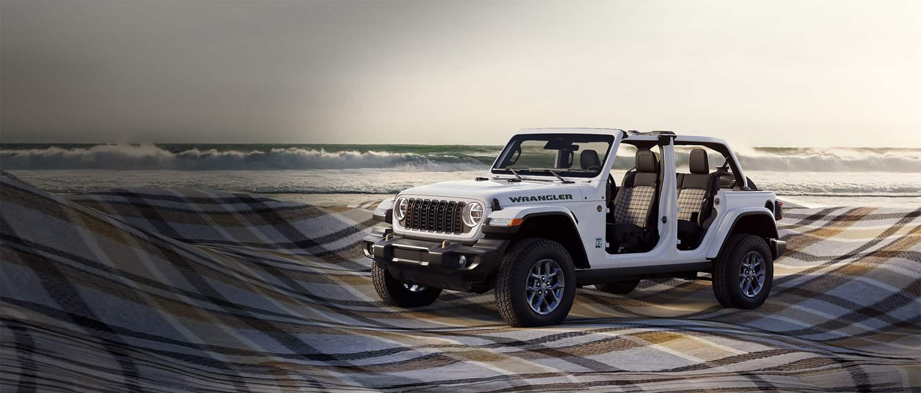 A driver-side front angle of a white 2026 Jeep Wrangler 85th Anniversary Edition with its doors and top removed, revealing plaid inserts on the front and rear seats. It is parked beside the ocean on a massive billowing plaid blanket.