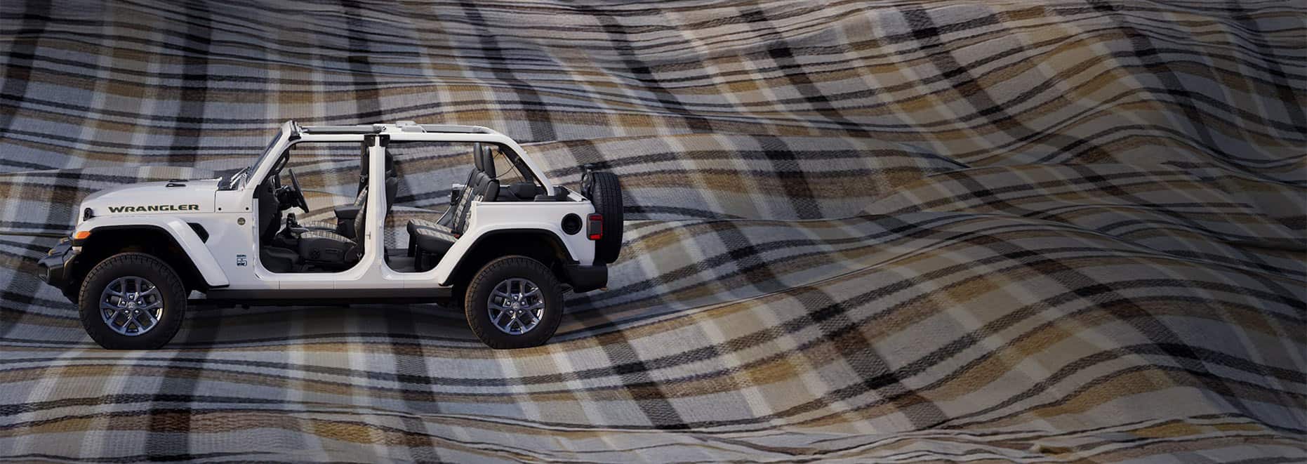 A driver-side profile of a white 2026 Jeep Wrangler 85th Anniversary Edition with its doors and top removed, revealing plaid inserts on the front and rear seats. It is parked on a massive billowing plaid blanket.