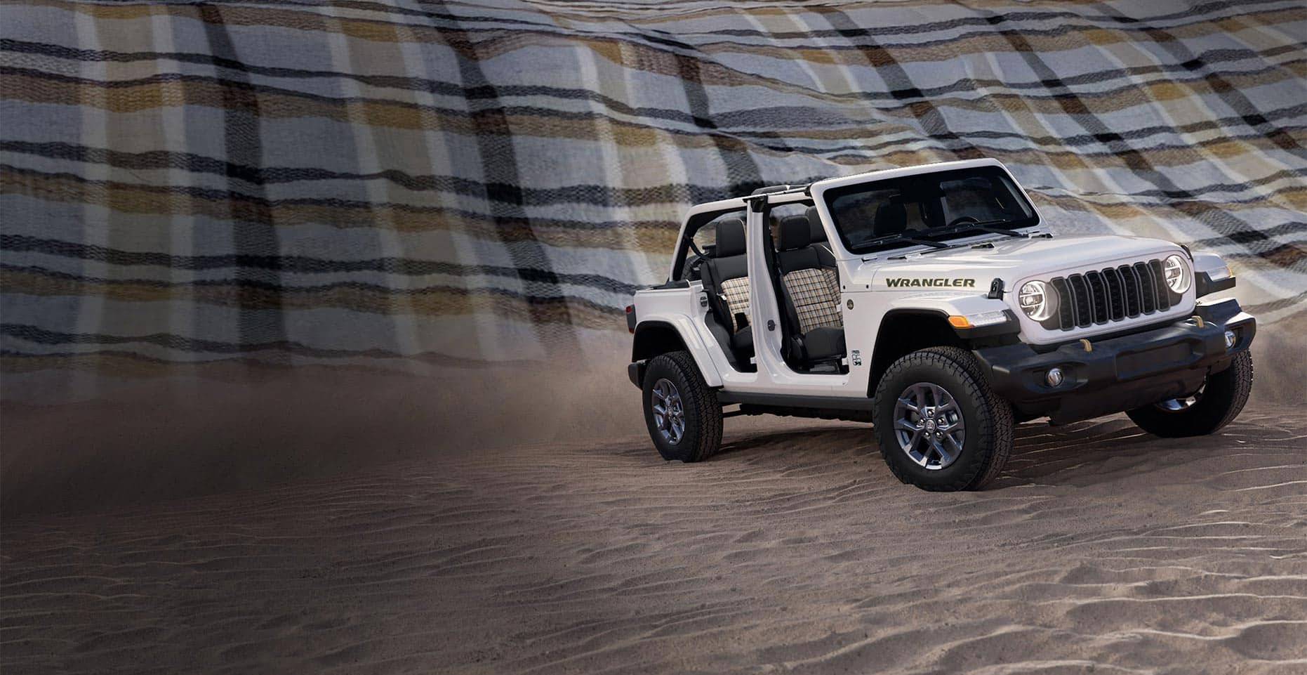 A passenger-side front angle of a white 2026 Jeep Wrangler 85th Anniversary Edition with its doors and top removed, revealing plaid inserts on the front and rear seats. It is parked on sand, with a massive plaid blanket as a backdrop.