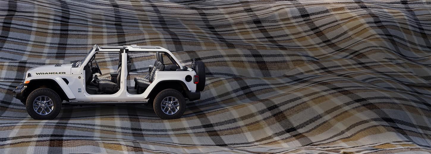 A driver-side profile of a white 2026 Jeep Wrangler 85th Anniversary Edition with its doors and top removed, revealing plaid inserts on the front and rear seats. It is parked on a massive billowing plaid blanket.