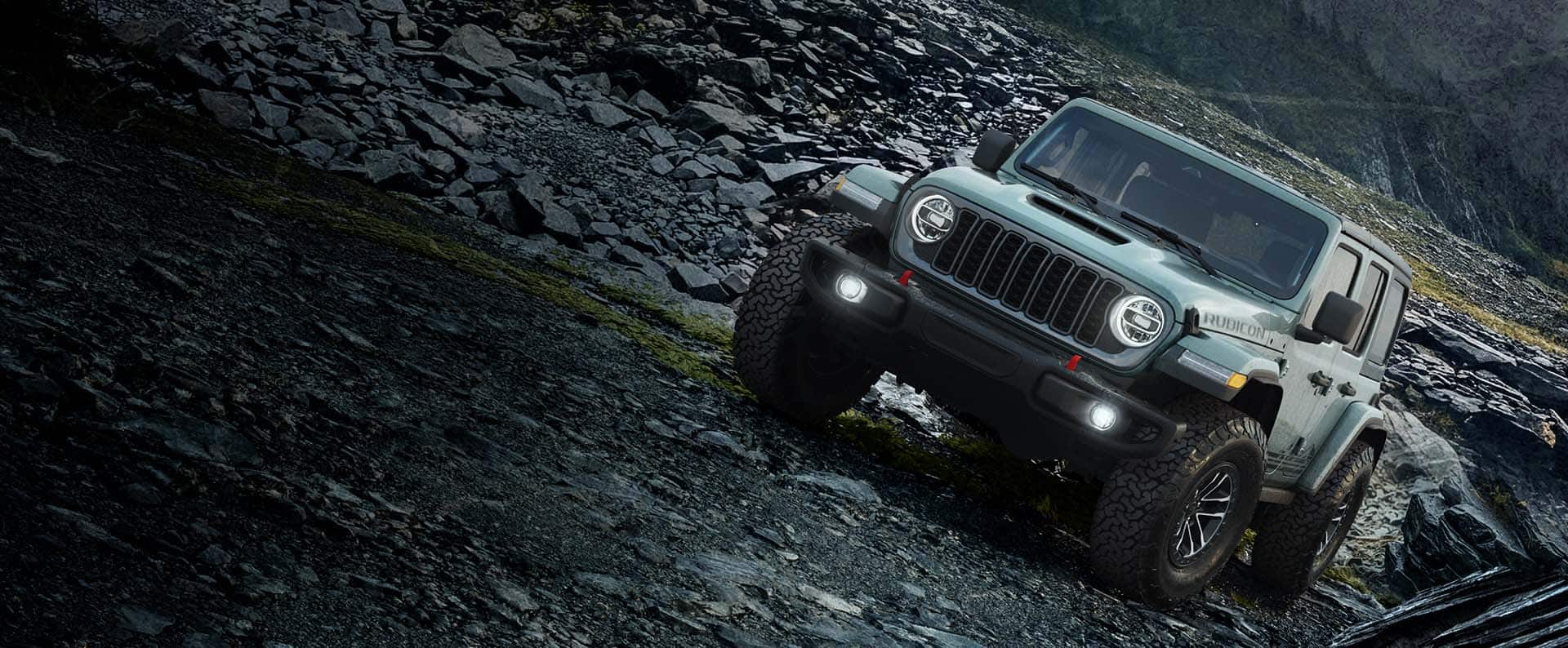 A front angle of a blue-green 2026 Jeep Wrangler Rubicon Rockslide crawling horizontally over a steep, rocky trail off-road.