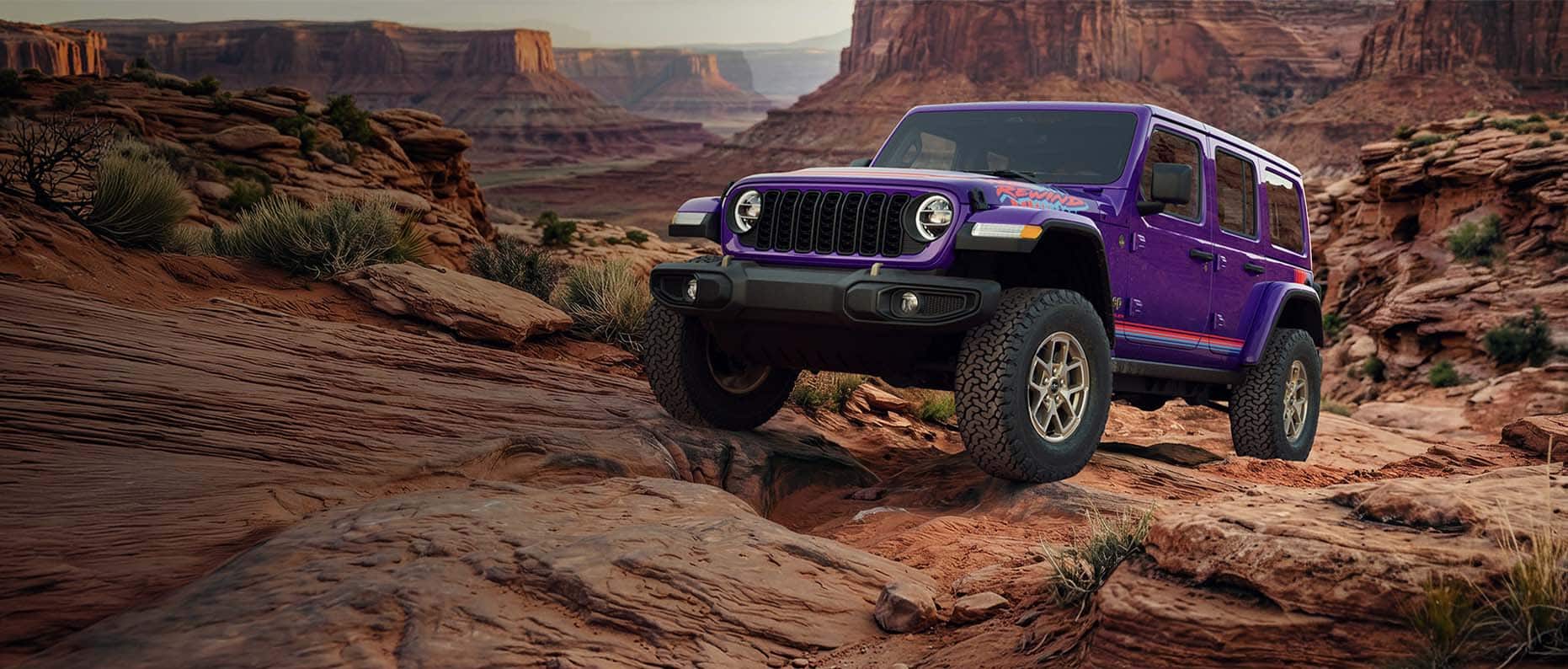 A purple 2026 Jeep Wrangler Rewind four-door with red and blue hood, fender and door decals, ascending a rocky hill off-road in the mountains.