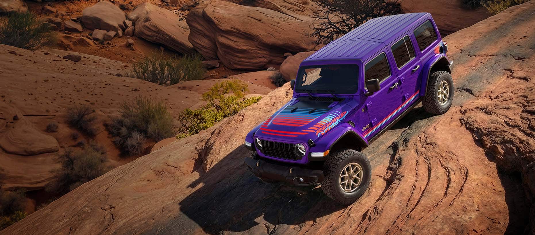 A purple 2026 Jeep Wrangler Rewind four-door with red and blue hood, fender and door decals, descending a very steep, rocky hill in the mountains.