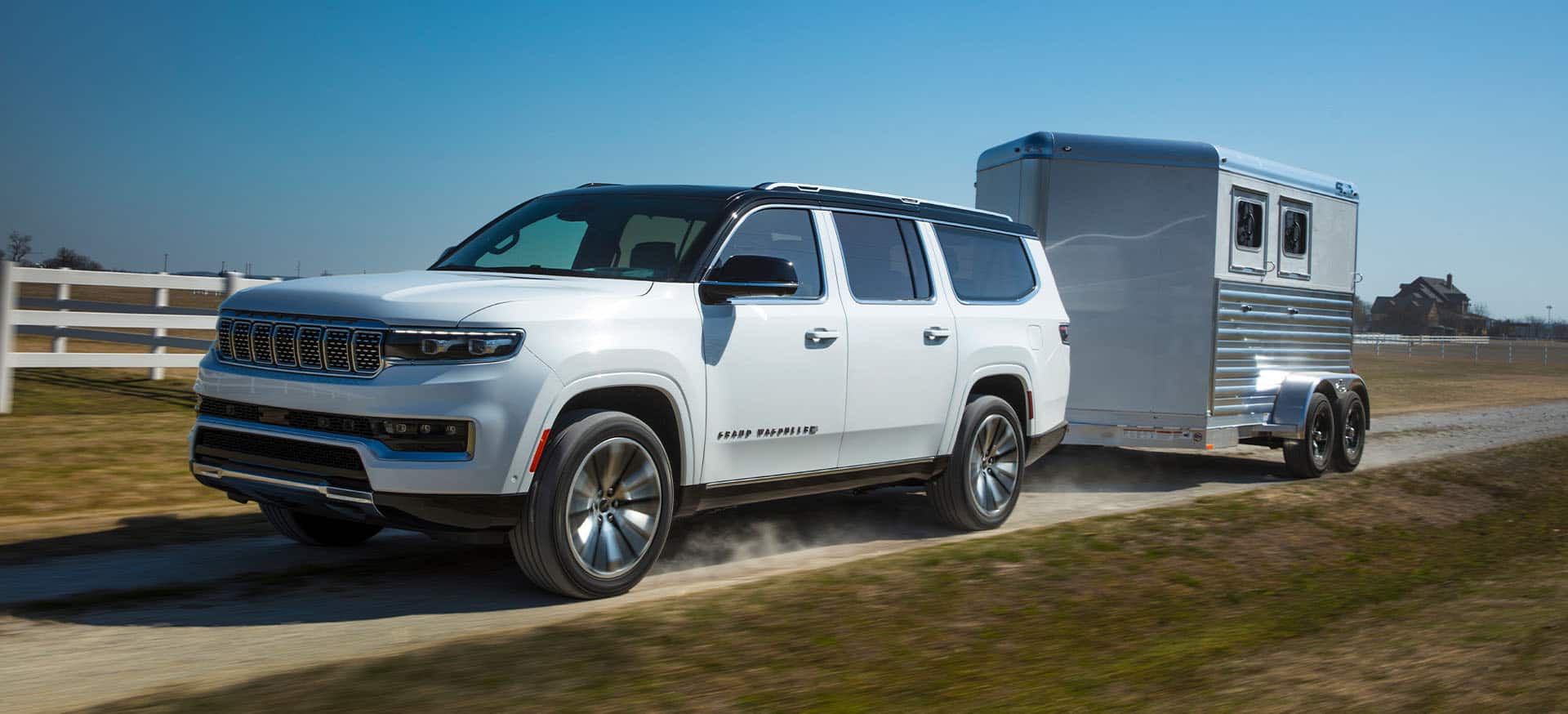 A white 2025 Jeep Grand Wagoneer Series 3 traveling down a dirt road beside a farm, towing a horse trailer.