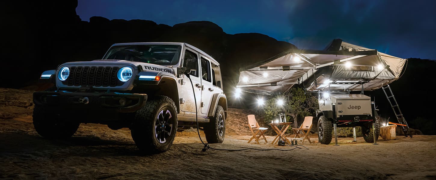 A 2025 Jeep Wrangler Rubicon X 4xe parked off-road in the desert at night with one end of a power cord plugged into its power box and next to camper trailer.