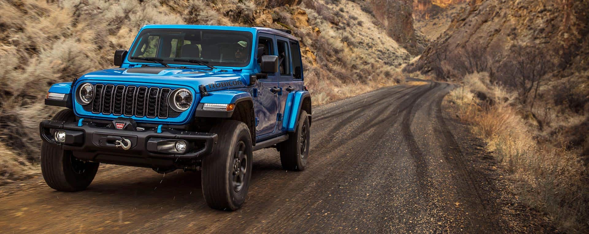 A blue 2024 Jeep Wrangler Rubicon 4xe being driven on a trail cut through the mountains.