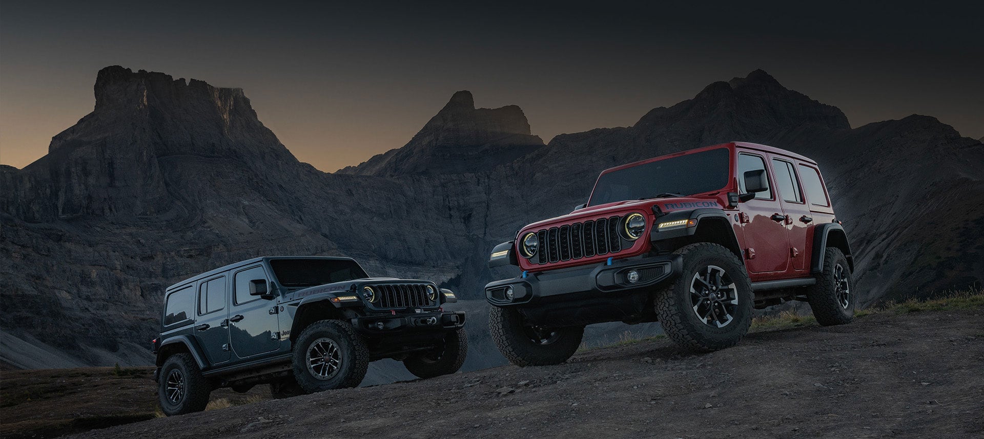 Two Jeep Wrangler models parked on a hill off-road at twilight: On the left, a blue 2025 Jeep Wrangler Rubicon X and on the right, a red 2025 Jeep Wrangler Rubicon 4xe.