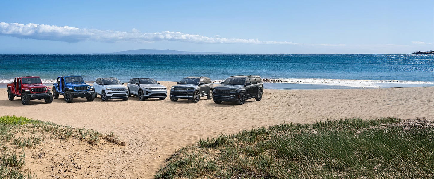 A lineup of six 2025 Jeep Brand vehicles parked on a sandy beach beside the water. From left to right: a red Gladiator Mojave X and blue Wrangler Rubicon X 4xe--both with their doors removed, a white Compass Limited, a silver Wagoneer S Launch Edition, a gray Grand Cherokee Summit Reserve and a black Grand Wagoneer Obsidian.